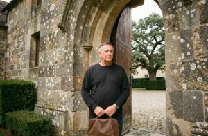 Un homme d'âge mûr se tenant à l'entrée d'un monastère français en pierre, tenant un sac en cuir, prêt à entrer dans la vie monastique